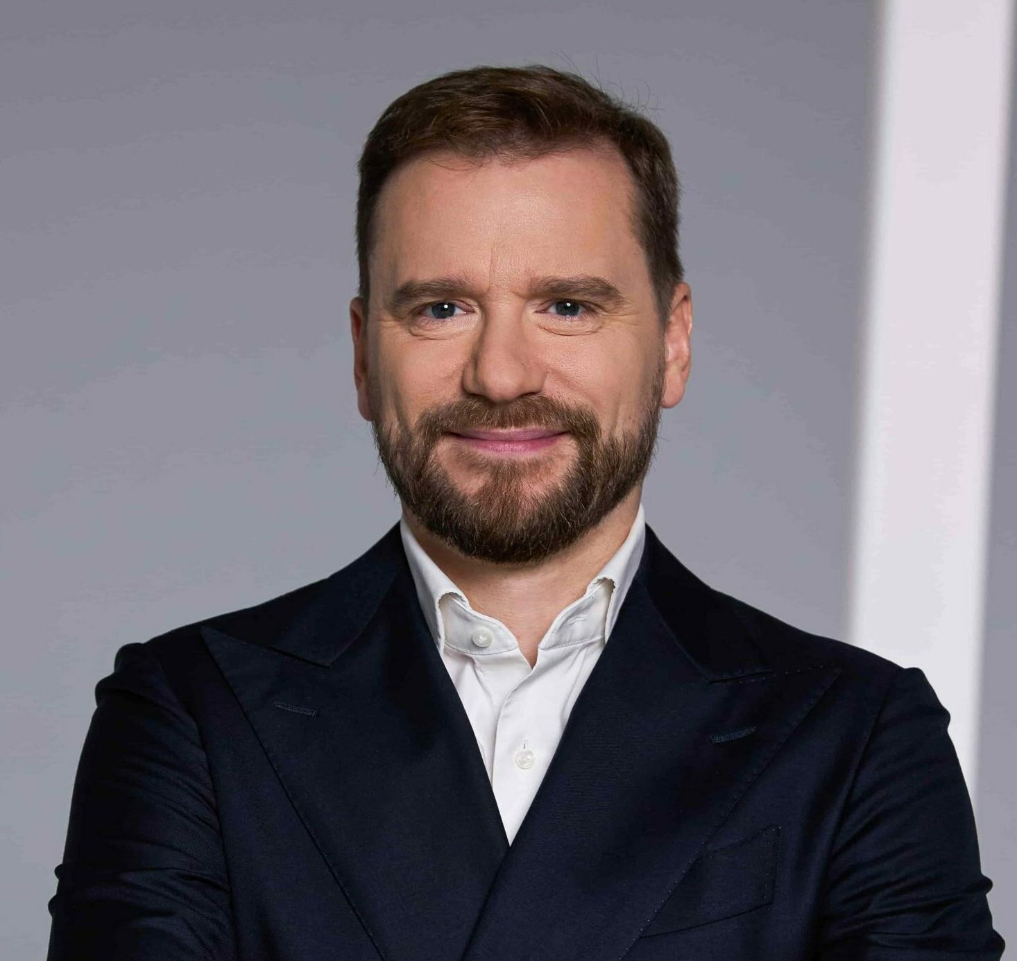 Donatas Frejus, dressed in a dark suit, standing with arms crossed and smiling in front of a light background