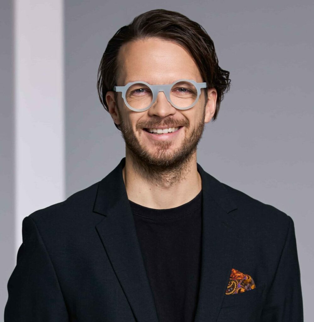 Benas Poderis, co-founder of ROIX, smiling in a dark blazer with glasses against a gray background